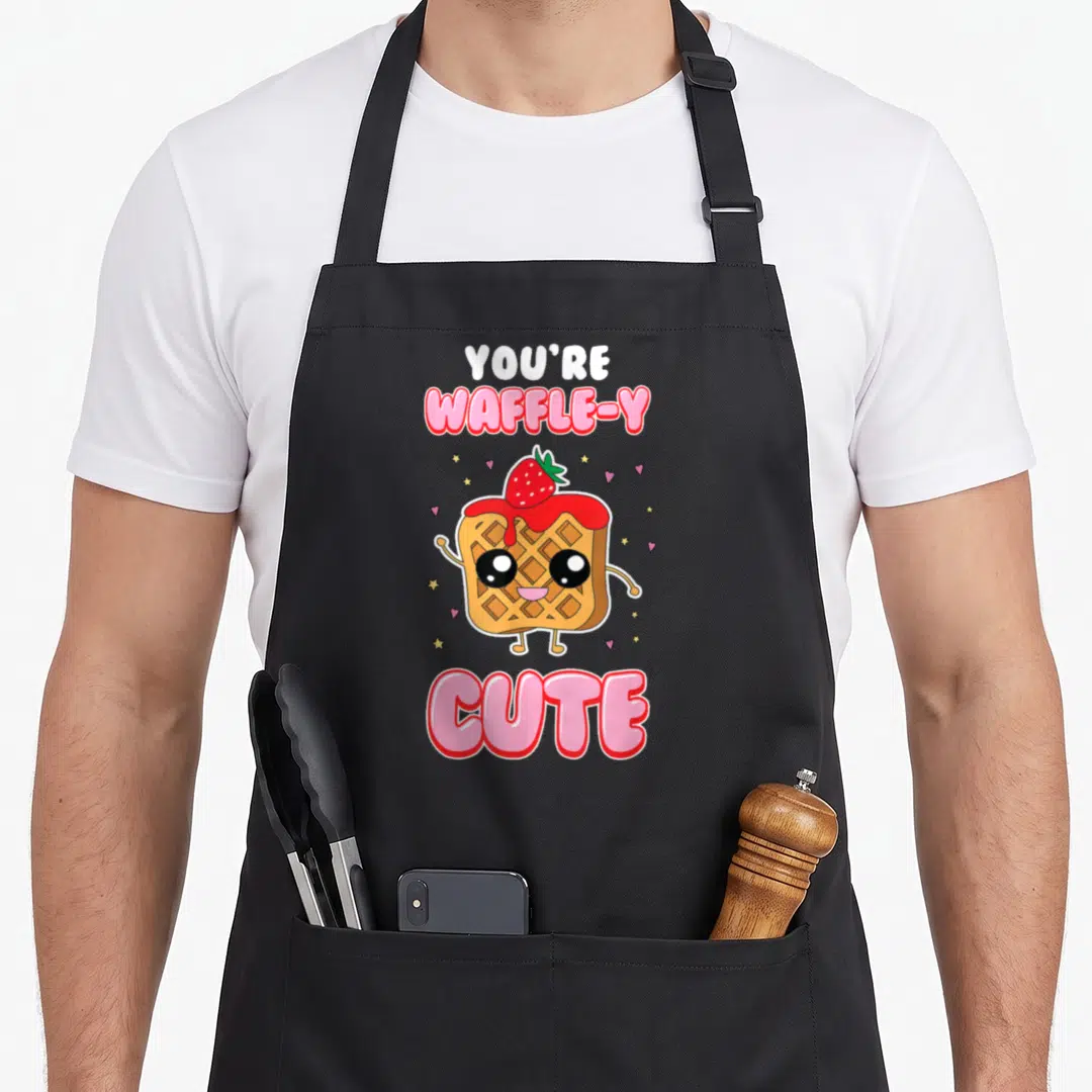 Awfully waffles apron - Cartoon waffle with syrup strawberry and text Youre Waffle-y Cute surrounded by hearts — shown on a black cotton canvas apron against a white background
