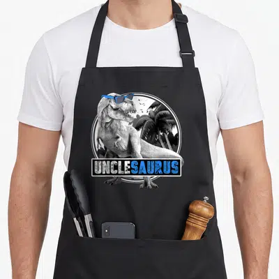 Unclesaurus rex dinosaur apron - T-Rex wearing blue sunglasses in front of palm trees with UNCLESAURUS text banner — shown on a black cotton canvas apron against a white background