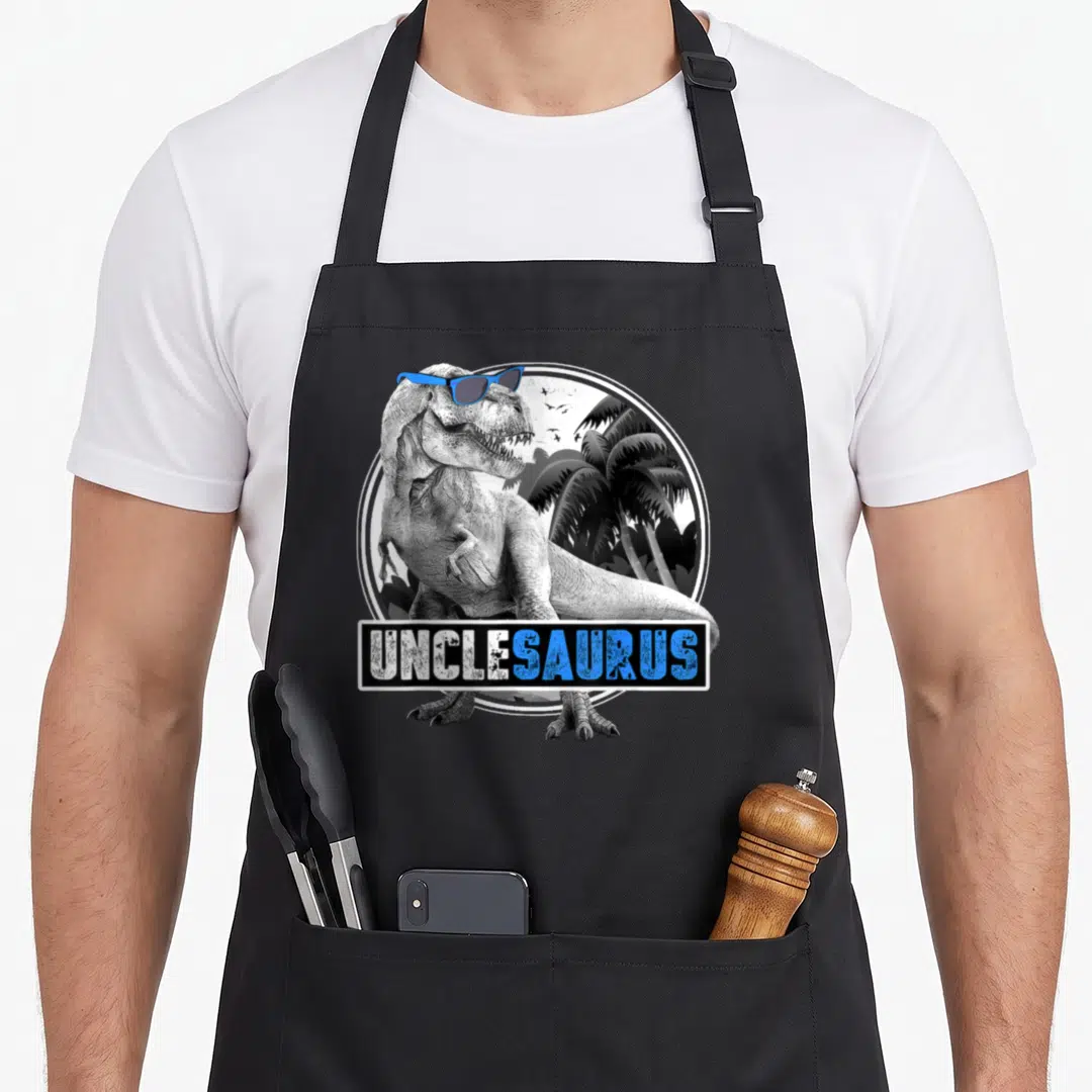 Unclesaurus rex dinosaur apron - T-Rex wearing blue sunglasses in front of palm trees with UNCLESAURUS text banner — shown on a black cotton canvas apron against a white background