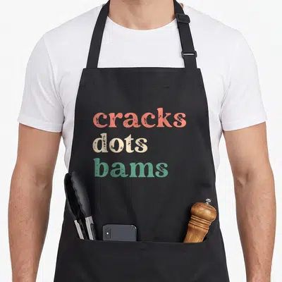 Dot mahjong apron - Stacked text reading cracks dots bams in coral beige and green distressed typography — shown on a black cotton canvas apron against a white background