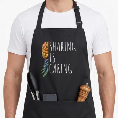 Sharing pineapple apron - Half pineapple in watercolor style beside stacked white text reading SHARING IS CARING — shown on a black cotton canvas apron against a white background
