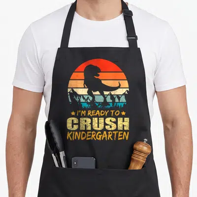 1st kindergarten dinosaur apron - Retro T-Rex silhouette with I'm Ready To Crush Kindergarten text and striped sun — shown on a black cotton canvas apron against a white background