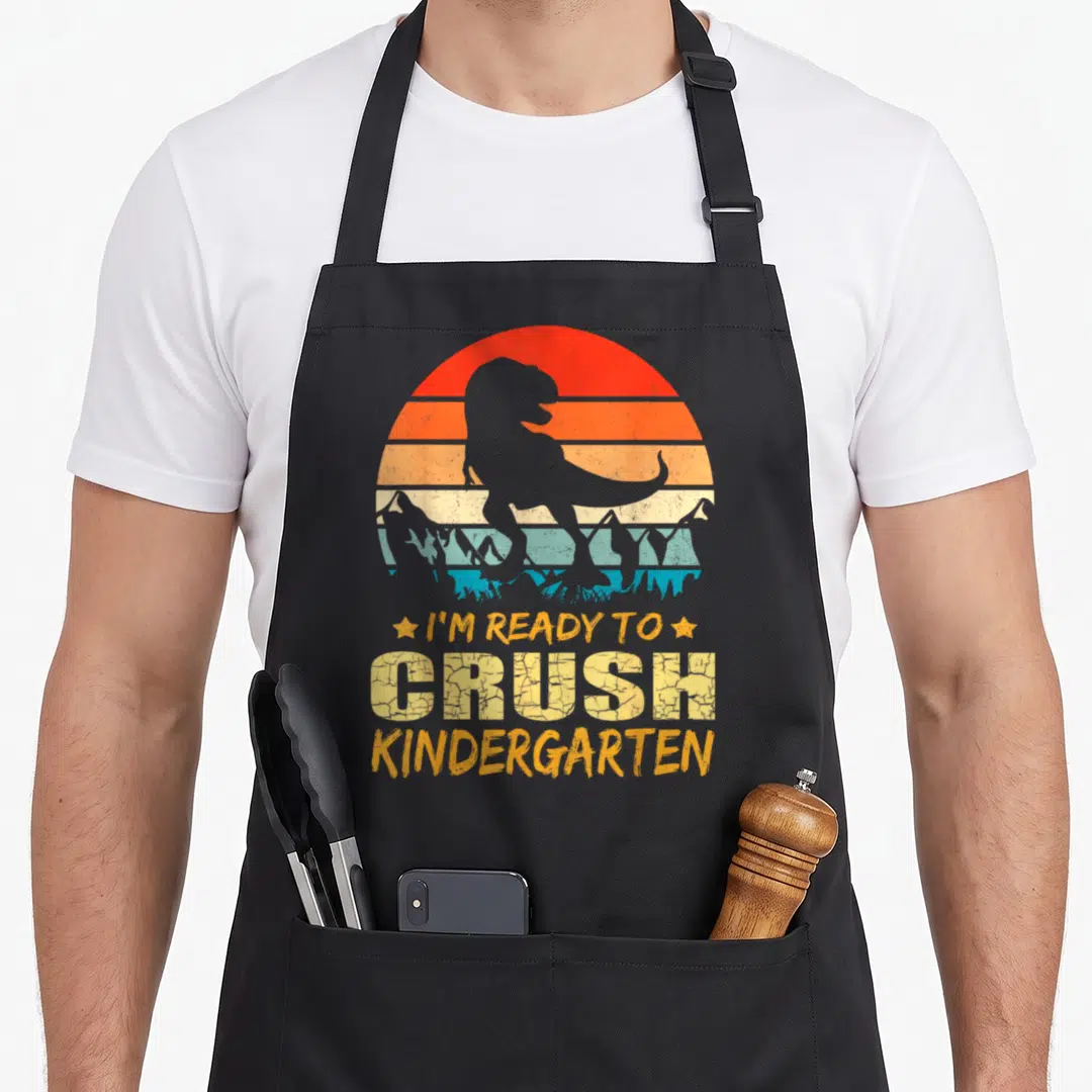 1st kindergarten dinosaur apron - Retro T-Rex silhouette with I'm Ready To Crush Kindergarten text and striped sun — shown on a black cotton canvas apron against a white background