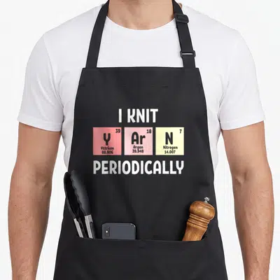 Scientist knitting apron - I KNIT Y Ar N PERIODICALLY text with Yttrium Argon Nitrogen colored squares — shown on a black cotton canvas apron against a white background