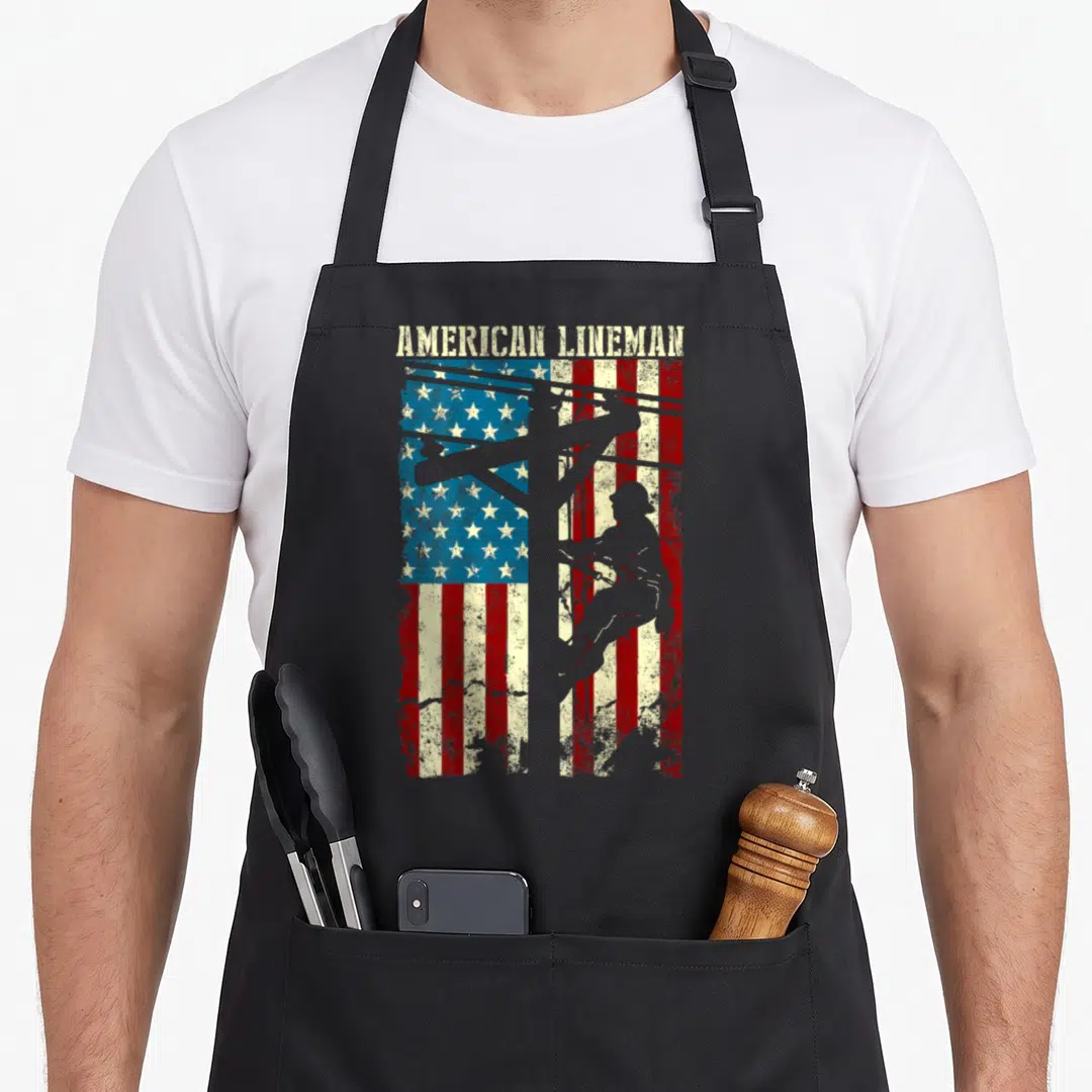 Lineman patriotic aprons - Distressed American flag with grey lineman silhouette climbing pole and AMERICAN LINEMAN text — shown on a black cotton canvas apron against a white background