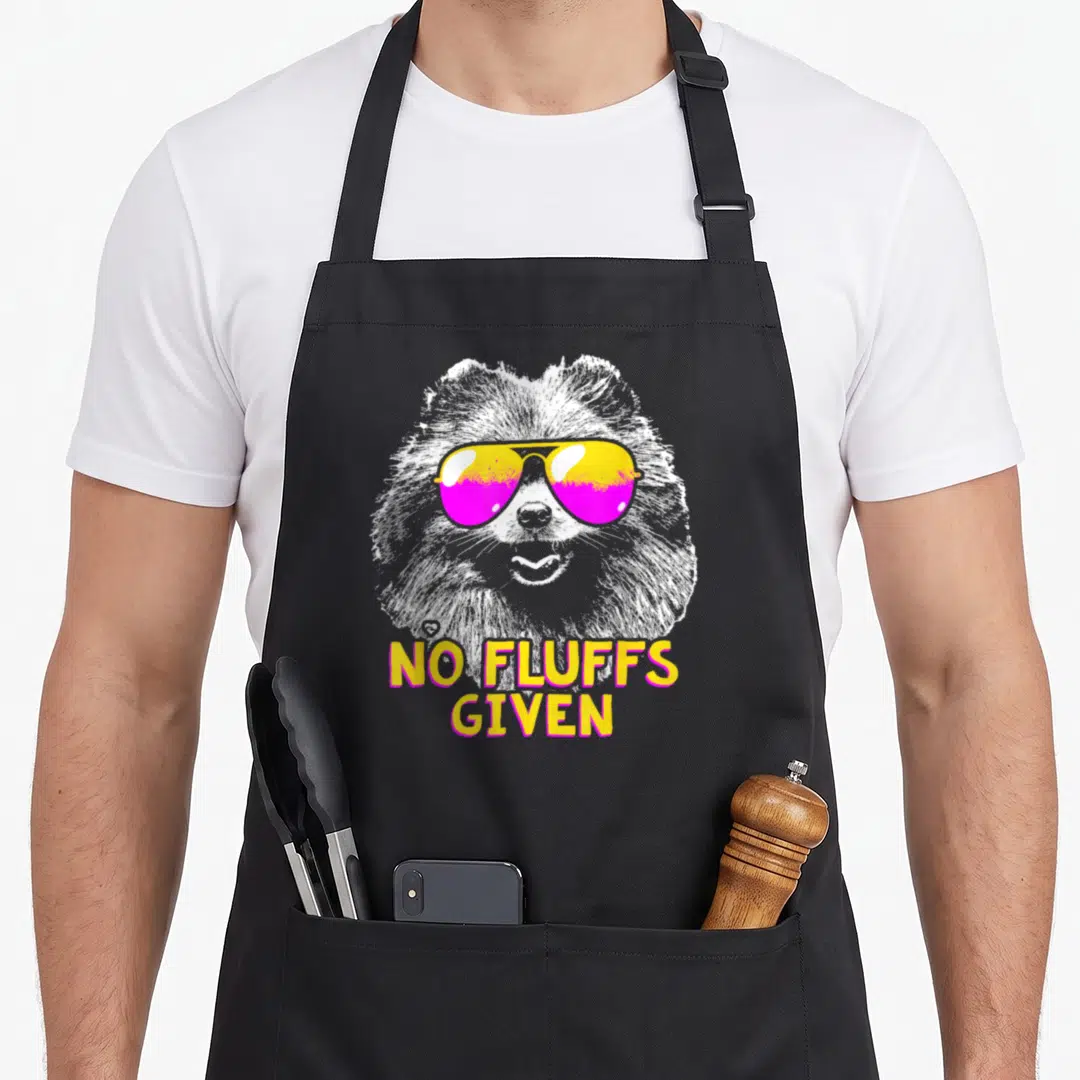 Fluff pomeranian apron - Cartoon white dog in yellow and pink sunglasses with NO FLUFFS GIVEN text — shown on a black cotton canvas apron against a white background