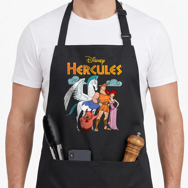 Hercule shot apron - Disney HERCULES text above cartoon hero, Megara, Phil, and Pegasus against light blue clouds — shown on a black cotton canvas apron against a white background