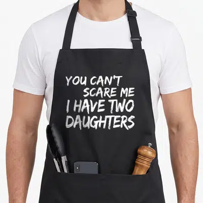 Retro scare father day apron - White stacked text reading YOU CAN'T SCARE ME I HAVE TWO DAUGHTERS in distressed font — shown on a black cotton canvas apron against a white background