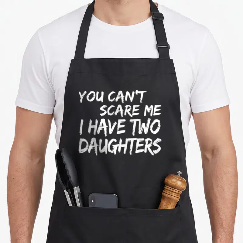 Retro scare father day apron - White stacked text reading YOU CAN'T SCARE ME I HAVE TWO DAUGHTERS in distressed font — shown on a black cotton canvas apron against a white background