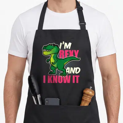 Youth dinosaur apron - Green cartoon T-Rex with sunglasses and text I'm Rexy And I Know It — shown on a black cotton canvas apron against a white background