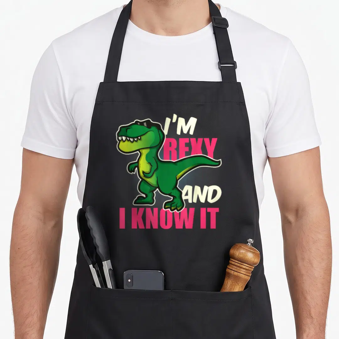 Youth dinosaur apron - Green cartoon T-Rex with sunglasses and text I'm Rexy And I Know It — shown on a black cotton canvas apron against a white background