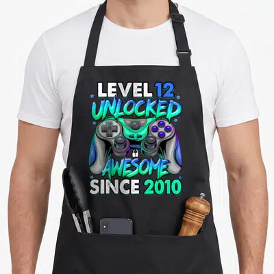 12th gaming aprons - Game controller with text LEVEL 12 UNLOCKED AWESOME SINCE 2010 in cyan green and blue cartoon style — shown on a black cotton canvas apron against a white background