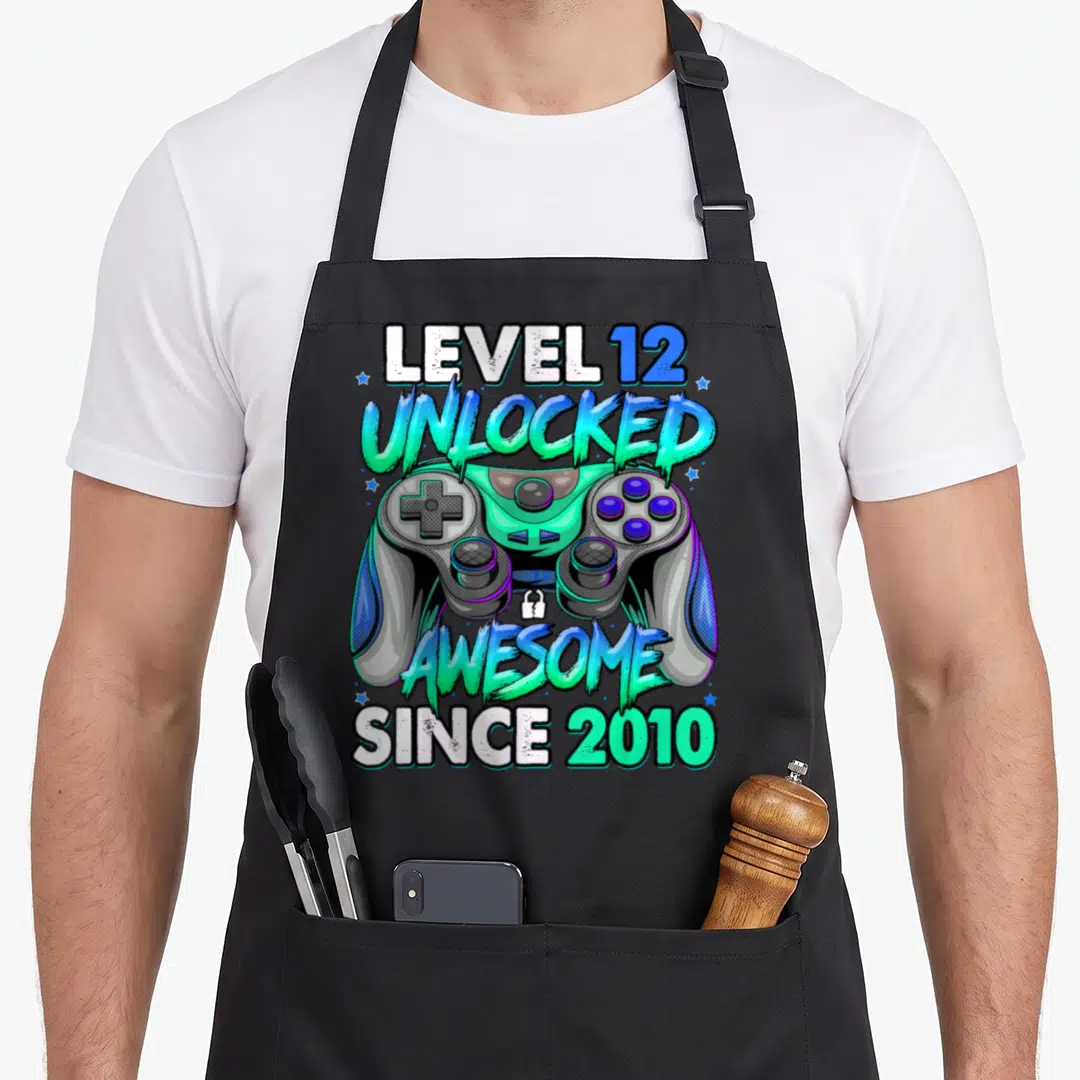 12th gaming aprons - Game controller with text LEVEL 12 UNLOCKED AWESOME SINCE 2010 in cyan green and blue cartoon style — shown on a black cotton canvas apron against a white background