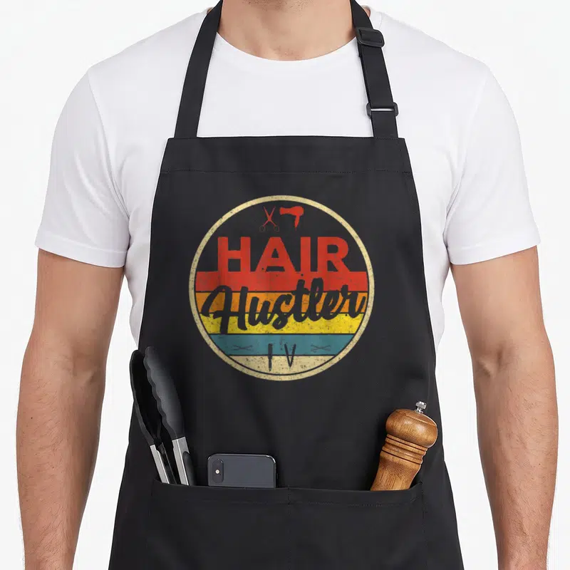 Hair hustler hairstylist apron - Retro circular graphic with HAIR Hustler IV text over colored stripes and red scissors — shown on a black cotton canvas apron against a white background