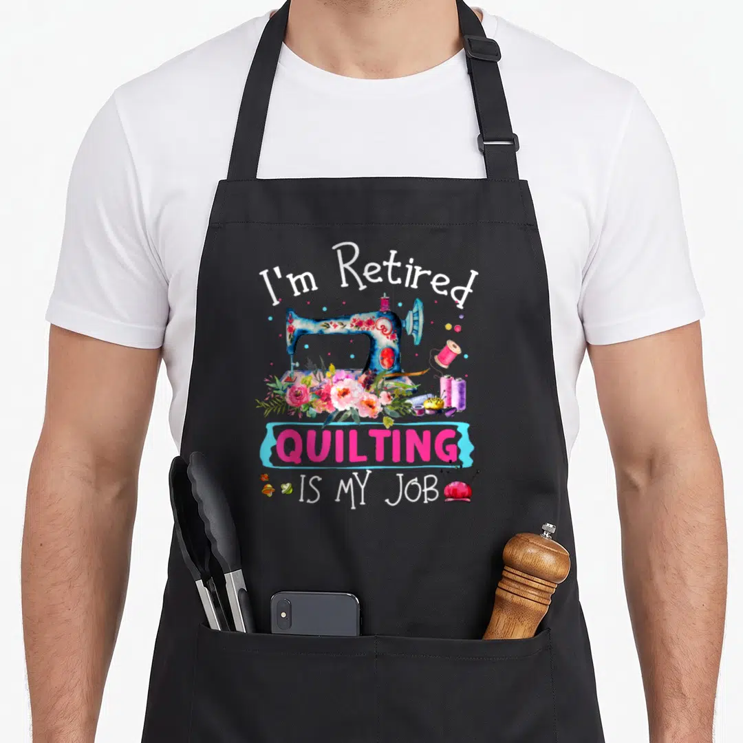Retired quilting apron - Floral sewing machine with I'm Retired QUILTING IS MY JOB text and pincushion — shown on a black cotton canvas apron against a white background