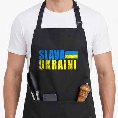 Slava distressed apron - Stacked blue and yellow text reading Slava Ukraini with a small rectangular flag beside — shown on a black cotton canvas apron against a white background