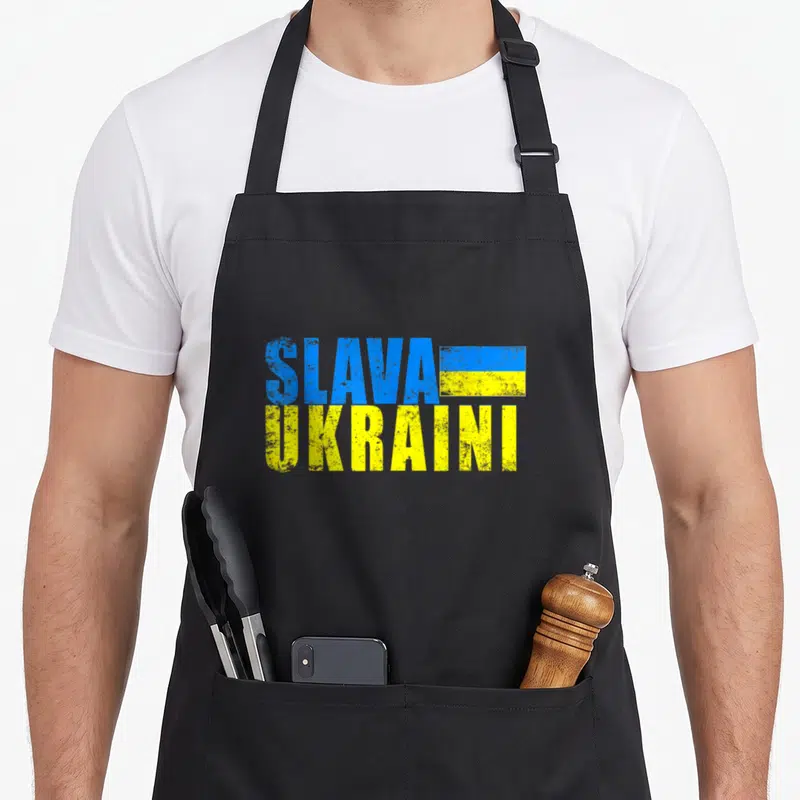Slava distressed apron - Stacked blue and yellow text reading Slava Ukraini with a small rectangular flag beside — shown on a black cotton canvas apron against a white background