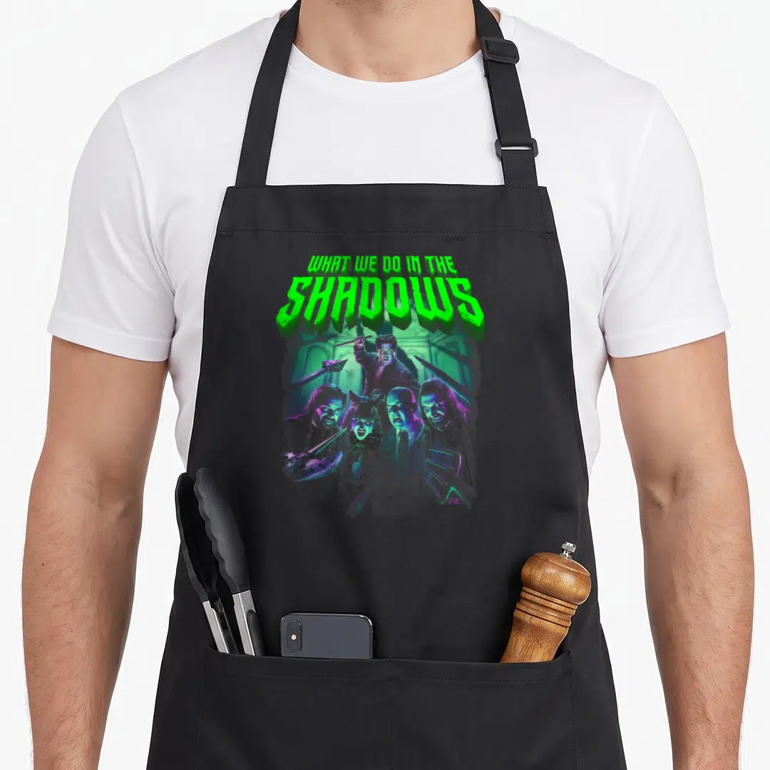 Shadow novelty apron - Green text WHAT WE DO IN THE SHADOWS above five armed vampires in purple and grey — shown on a black cotton canvas apron against a white background