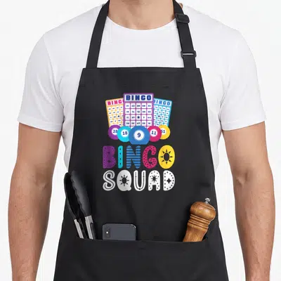 Humour bingo aprons - Three bingo cards and five chips above colorful BINGO SQUAD text in cartoon style — shown on a black cotton canvas apron against a white background