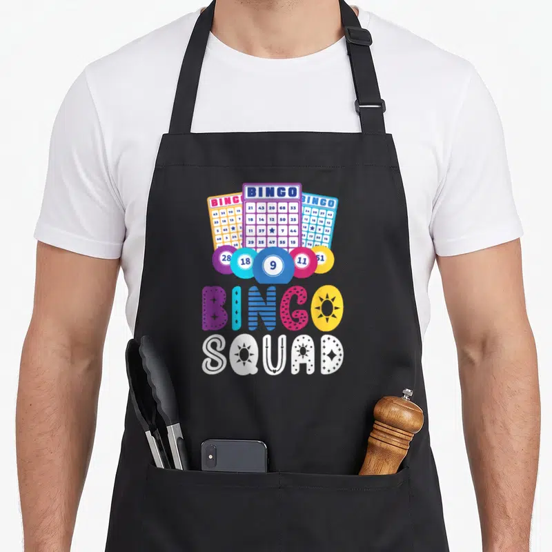 Humour bingo aprons - Three bingo cards and five chips above colorful BINGO SQUAD text in cartoon style — shown on a black cotton canvas apron against a white background