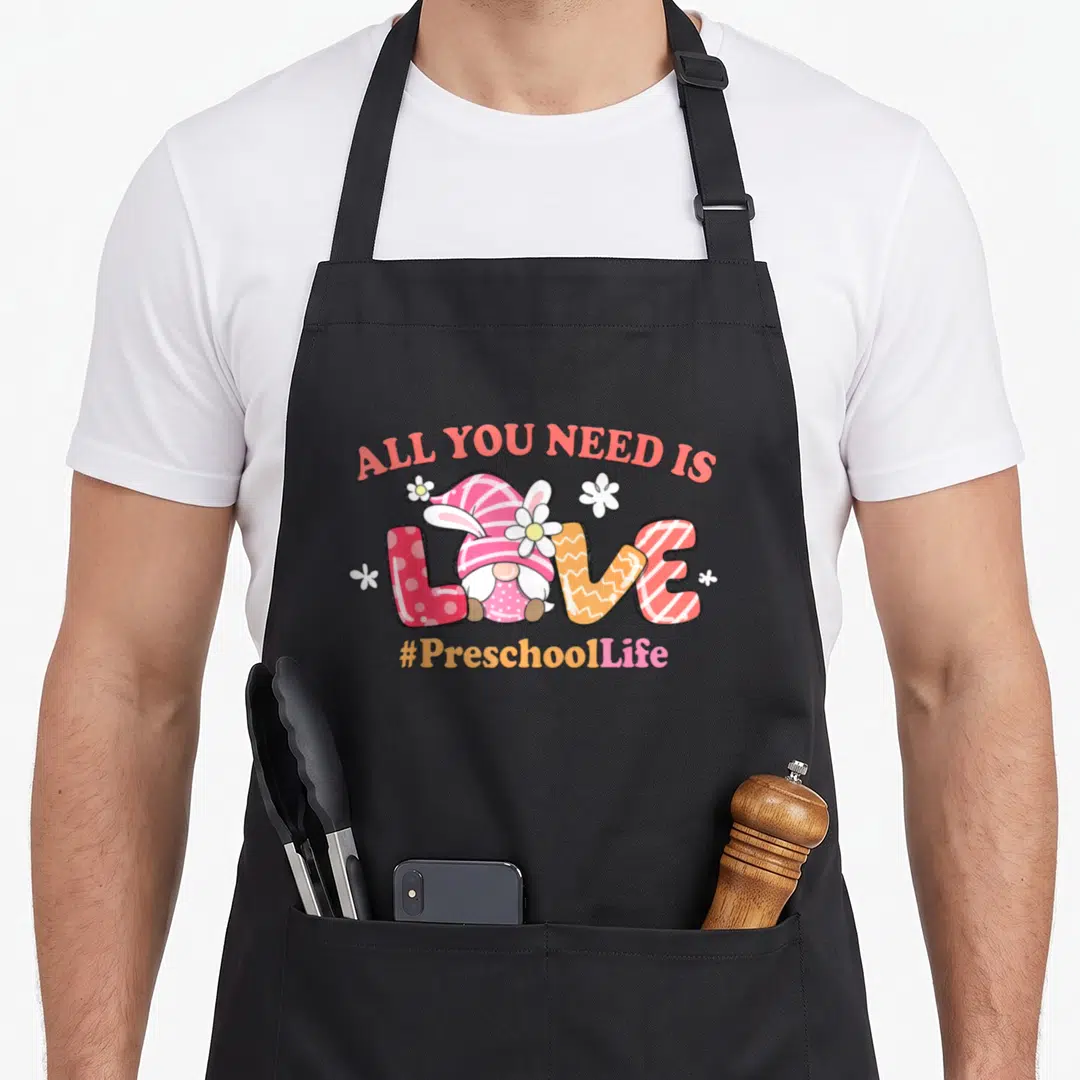 Gnome preschool teacher apron - Cartoon gnome sitting in LOVE text with ALL YOU NEED IS and #PreschoolLife surrounded — shown on a black cotton canvas apron against a white background