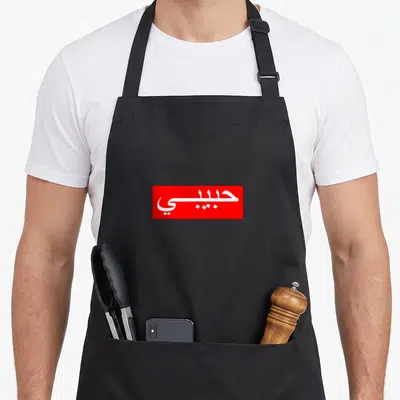 Habibi arab apron - White Arabic script reading Habibi inside a solid red rectangular block — shown on a black cotton canvas apron against a white background