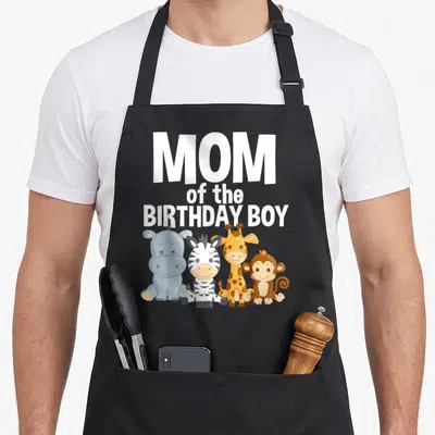 Safari zoo birthday apron - MOM of the BIRTHDAY BOY text above four cartoon animals including hippo, zebra, giraffe — shown on a black cotton canvas apron against a white background