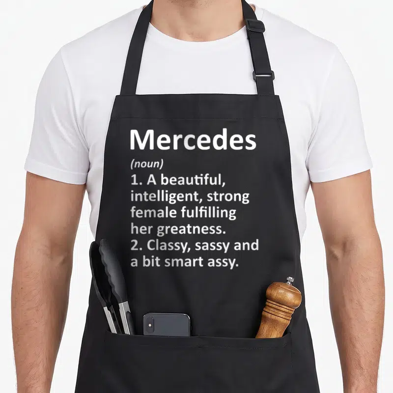 Meaning wife apron - White text defining Mercedes as a beautiful, intelligent, sassy female fulfilling her greatness — shown on a black cotton canvas apron against a white background