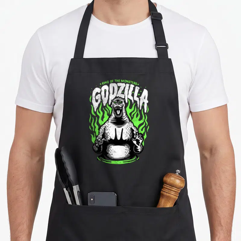 Movie flame apron - Roaring Godzilla figure surrounded by green flames with KING OF THE MONSTERS text above — shown on a black cotton canvas apron against a white background