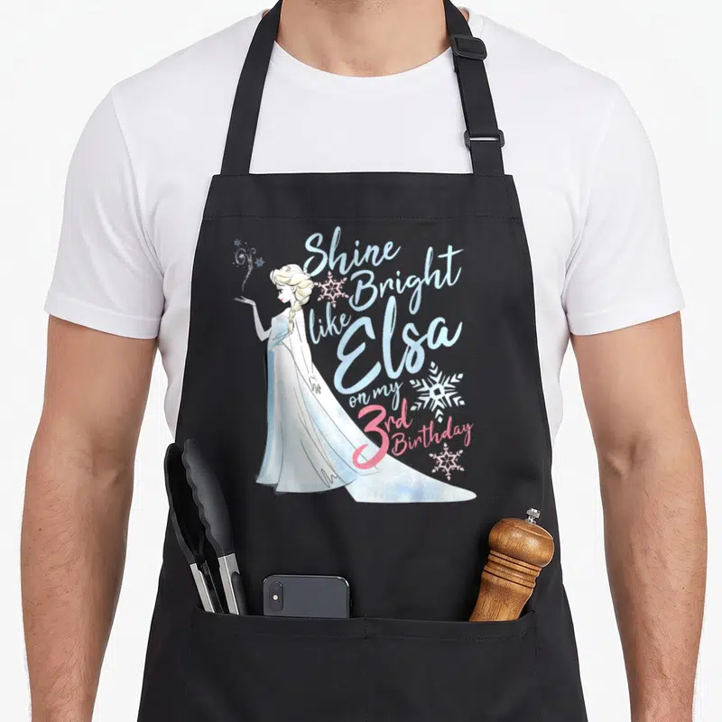 Shine bright birthday apron - Elsa standing next to stacked text reading Shine Bright like Elsa on my 3rd Birthday — shown on a black cotton canvas apron against a white background