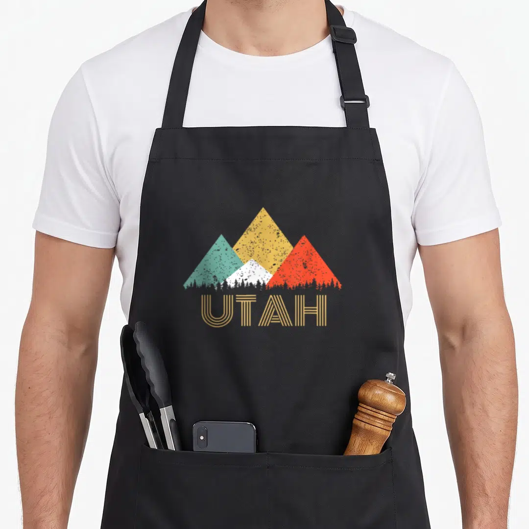 Mountain utah pride apron - Four geometric mountains in teal yellow red and white above UTAH text and tree line — shown on a black cotton canvas apron against a white background