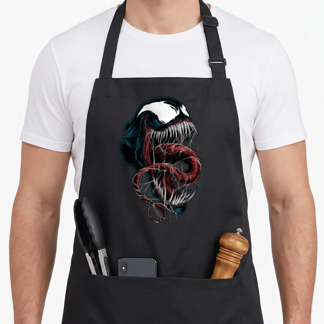 Comic face apron - Realistic Venom head with open mouth and curling tongue in black, red, white, and blue — shown on a black cotton canvas apron against a white background