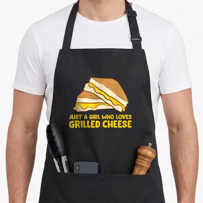 Cheese grilled apron - Cartoon stacked grilled cheese sandwich with yellow text reading JUST A GIRL WHO LOVES GRILLED — shown on a black cotton canvas apron against a white background