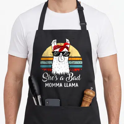 Momma mama apron - Cartoon white llama with red bandana and sunglasses in front of striped sun and white text — shown on a black cotton canvas apron against a white background