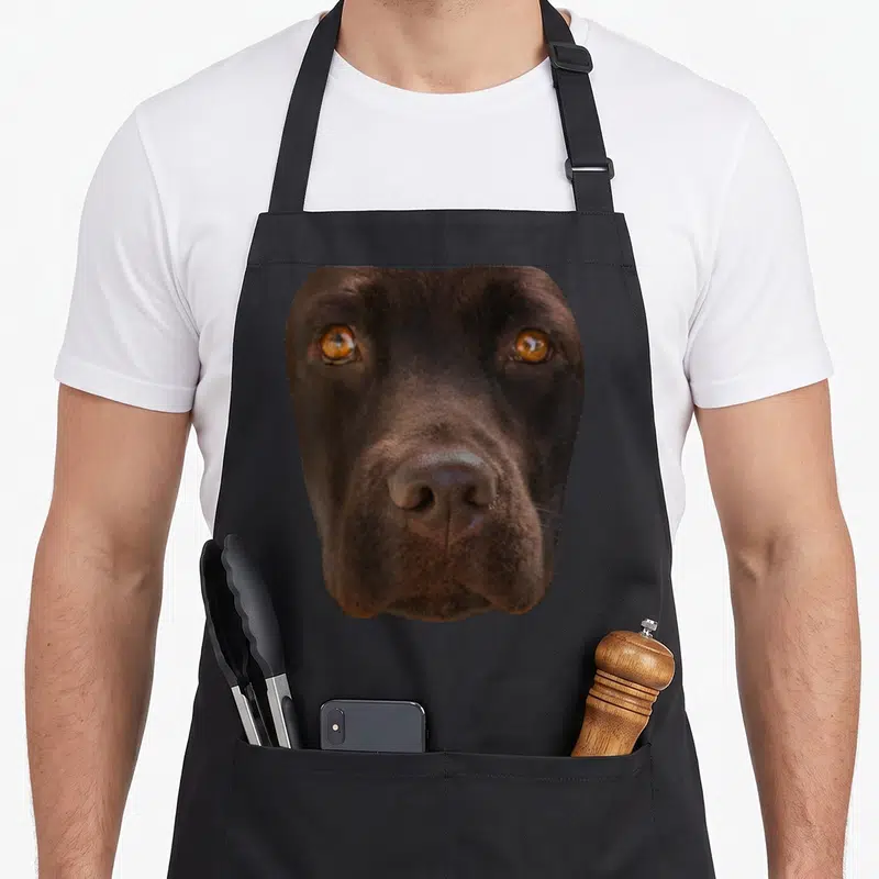 Chocolate dark brown apron - Realistic dog face photo print in brown and amber fur tones — shown on a black cotton canvas apron against a white background