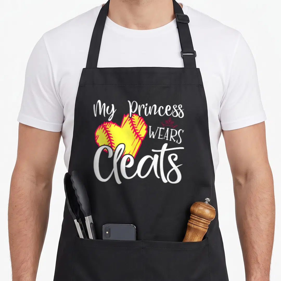 Cleat princess apron - My Princess Wears Cleats text around yellow heart softball with red stitching and crown — shown on a black cotton canvas apron against a white background