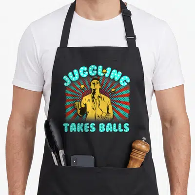 Juggler juggling apron - Retro juggler figure with JUGGLING TAKES BALLS text on red and blue sunburst — shown on a black cotton canvas apron against a white background