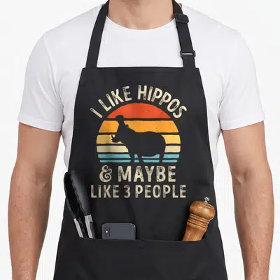 Maybe hippopotamus apron - I LIKE HIPPOS text over retro striped sunset with grey hippo silhouette — shown on a black cotton canvas apron against a white background