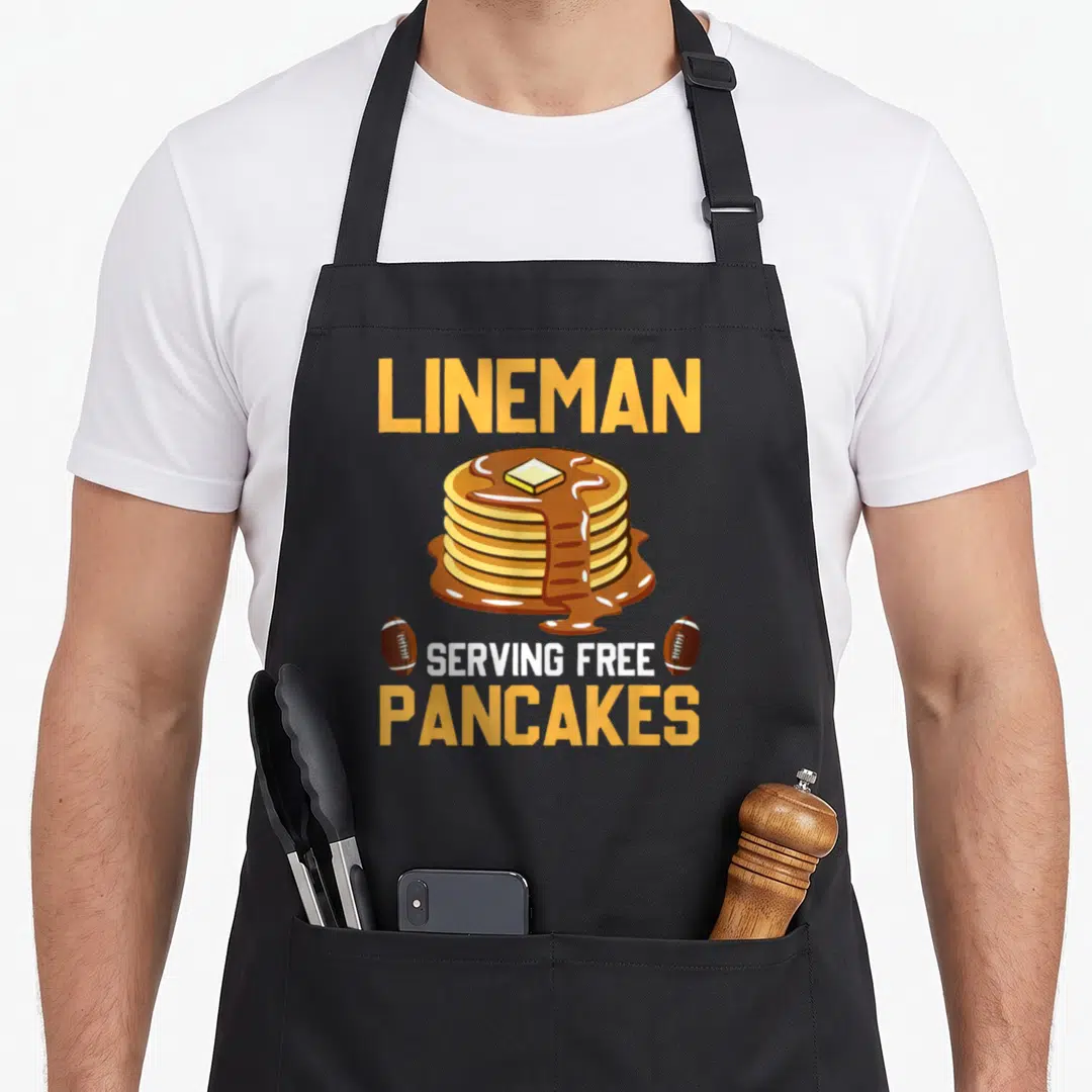 Serve pancake apron - Cartoon pancakes stack with butter and two footballs under LINEMAN text — shown on a black cotton canvas apron against a white background