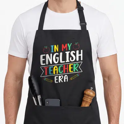 Era english chef apron - IN MY ENGLISH TEACHER ERA text with two apples and two pencils in cartoon style — shown on a black cotton canvas apron against a white background