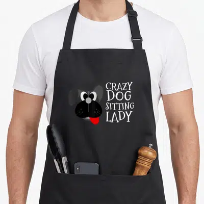 Sitter dog cooking apron - Cartoon dog head with red tongue next to stacked white text Crazy Dog Sitting Lady — shown on a black cotton canvas apron against a white background