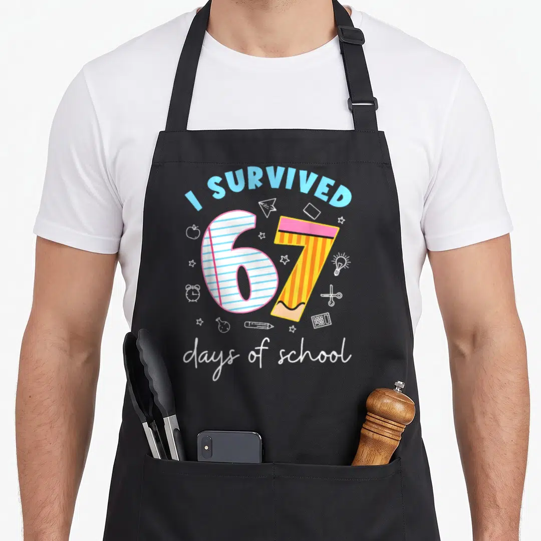 Meme teacher apron - I Survived 67 Days of School graphic with pencil number 7 and lined number 6 surrounded by school — shown on a black cotton canvas apron against a white background