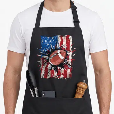 Patriotic football 4th of july apron - Brown football over distressed USA flag with red white blue and black splatter — shown on a black cotton canvas apron against a white background