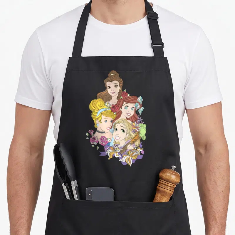 Floral princess belle apron - Four cartoon princesses clustered with red roses and a violin — shown on a black cotton canvas apron against a white background