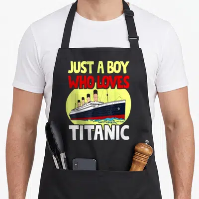 Titanic novelty apron - Cartoon ship in yellow circle with text Just a Boy Who Loves Titanic — shown on a black cotton canvas apron against a white background