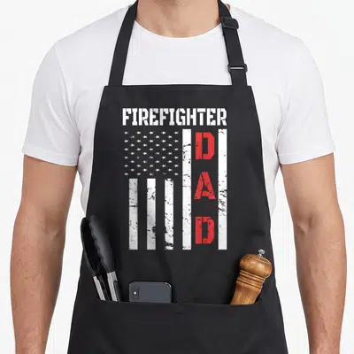 Flag father firefighter aprons - Distressed USA flag with vertical red stripes and stenciled FIREFIGHTER DAD text — shown on a black cotton canvas apron against a white background