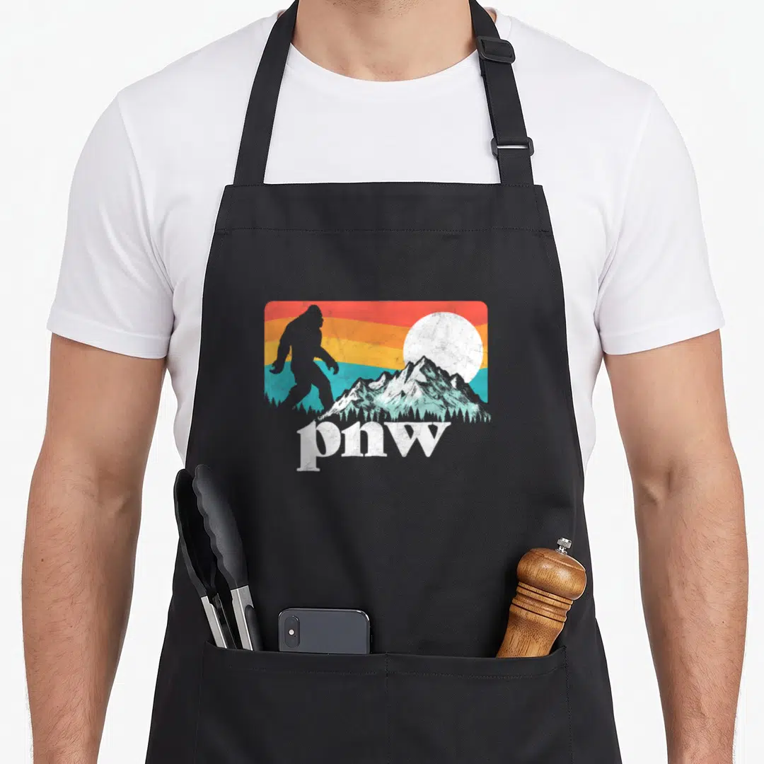 Pnw bigfoot apron - Retro Bigfoot silhouette against striped sunset with mountains and pnw text — shown on a black cotton canvas apron against a white background