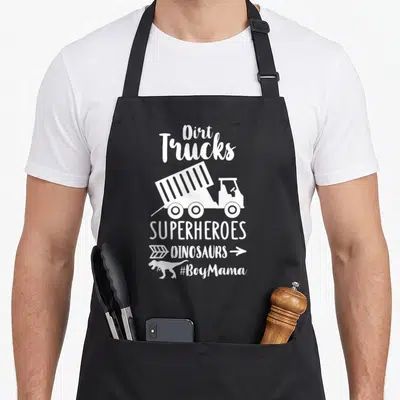 Superheroe truck apron - White dump truck and dinosaur silhouette with stacked text reading Dirt Trucks Superheroes — shown on a black cotton canvas apron against a white background