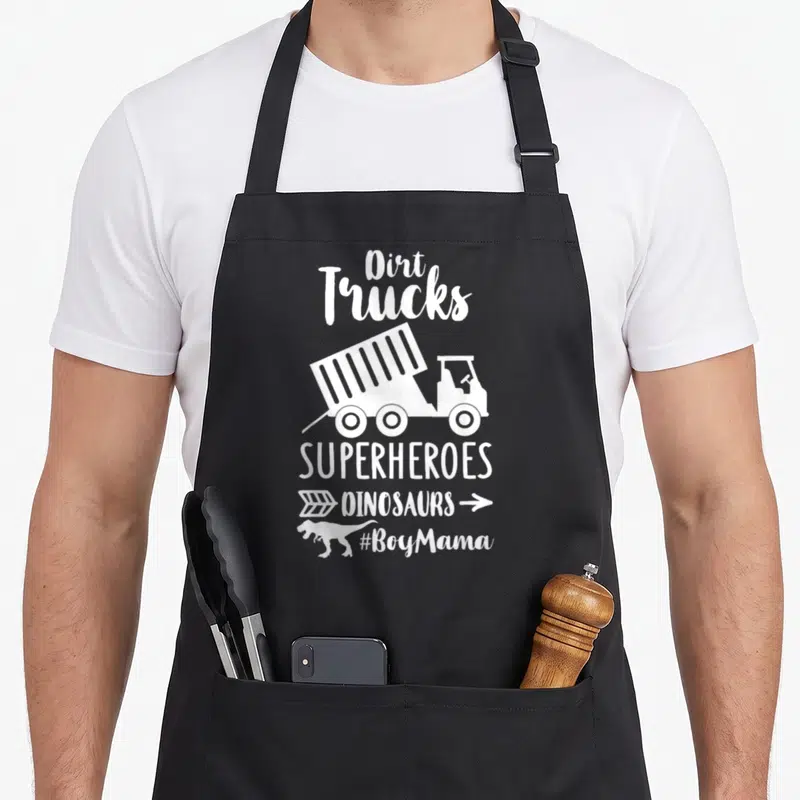 Superheroe truck apron - White dump truck and dinosaur silhouette with stacked text reading Dirt Trucks Superheroes — shown on a black cotton canvas apron against a white background