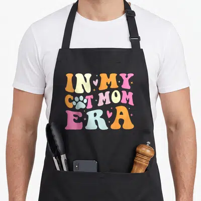 Funny era cat apron - Stacked retro text In My Cat Mom Era with paw print and pastel colors — shown on a black cotton canvas apron against a white background
